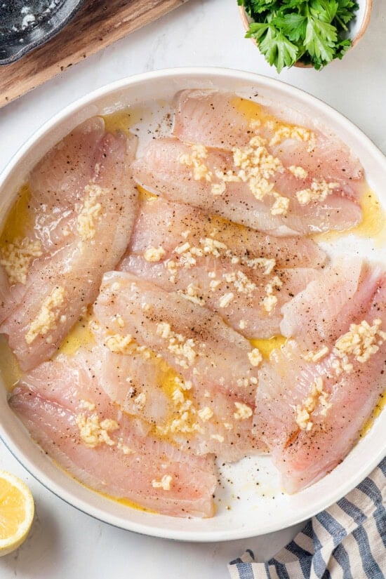 This simple baked tilapia recipe is finished with a garlicky lemon butter sauce and parsley, making it a delicious and light weeknight dinner! Overhead view of tilapia fillets in pan before baking
