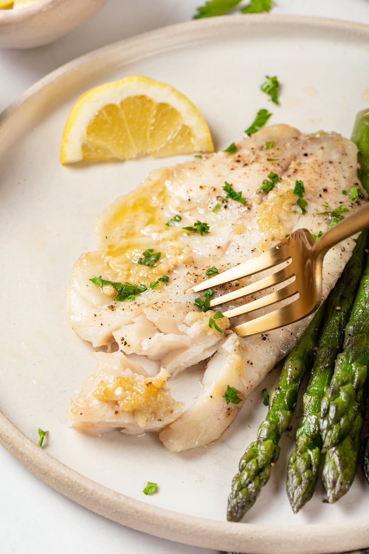 This simple baked tilapia recipe is finished with a garlicky lemon butter sauce and parsley, making it a delicious and light weeknight dinner! Fork cutting into baked tilapia on plate with asparagus