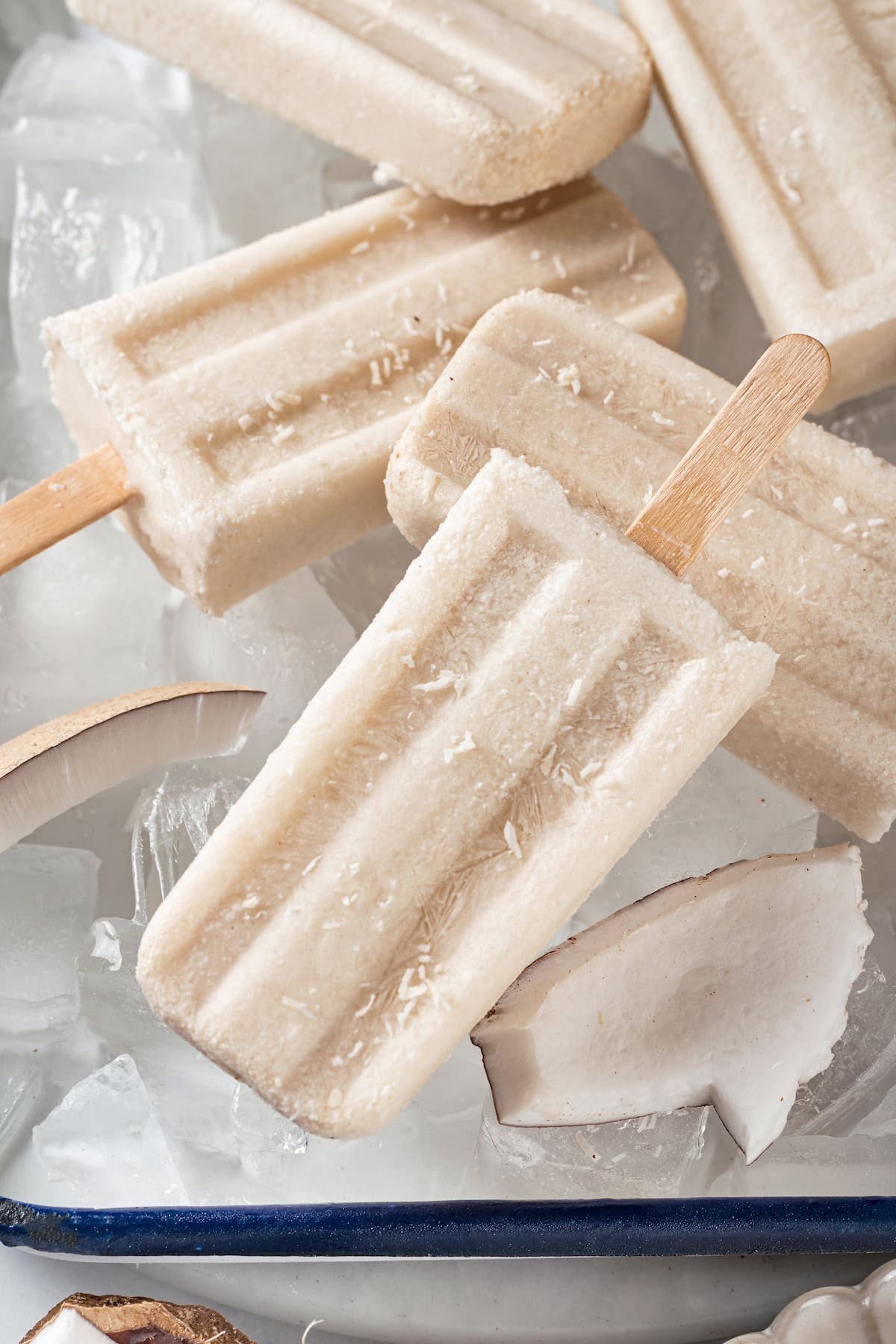 These homemade coconut popsicles are rich, creamy, and super simple to make! They’re the perfect summertime refresher. Homemade coconut popsicles on ice with coconut pieces