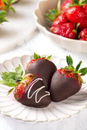 Three chocolate-covered strawberries on white plate with bowl of berries in background