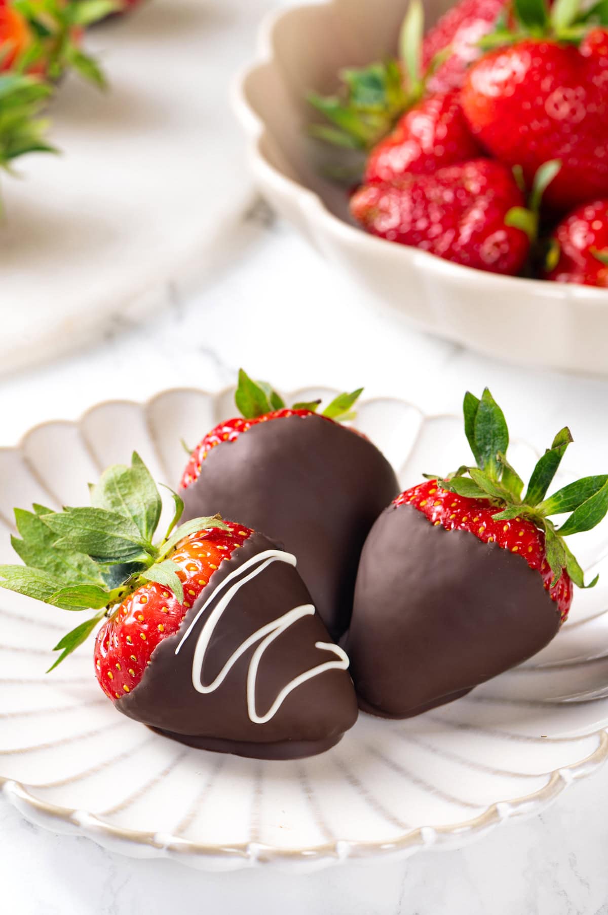 Chocolate-covered strawberries are an elegant treat that's incredibly easy to make at home. You'll never buy them at the store again! Three chocolate-covered strawberries on white plate with bowl of berries in background