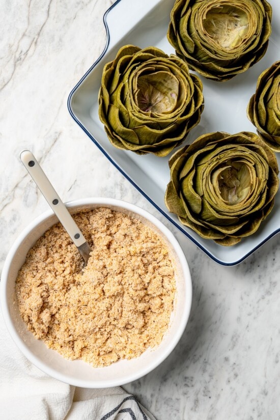 Stuffed artichokes are a classic Italian dish made with crispy, cheesy breadcrumbs. My version is a bit lighter but just as delicious! Overhead view of artichokes in baking dish and breadcrumb mixture in bowl