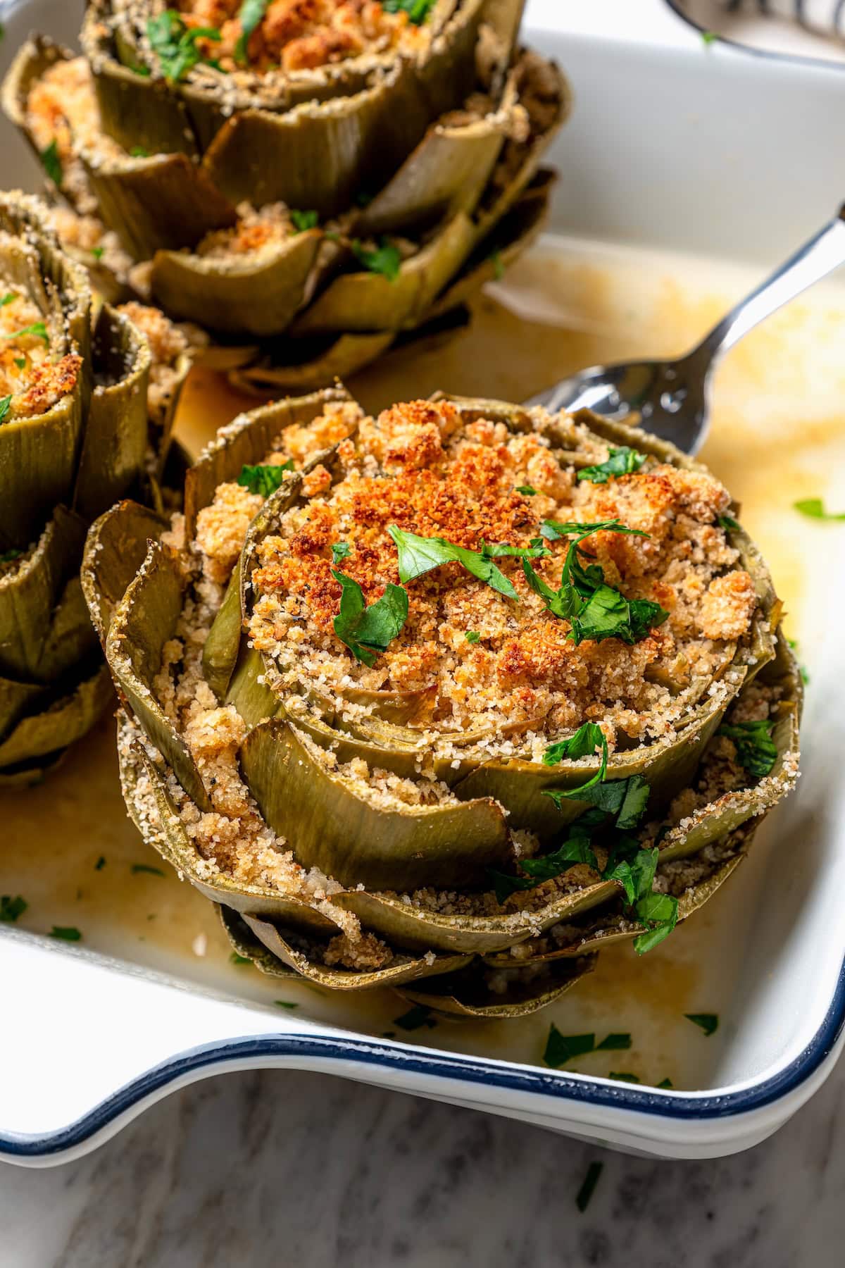 Stuffed artichokes are a classic Italian dish made with crispy, cheesy breadcrumbs. My version is a bit lighter but just as delicious! Fork lifting stuffed artichoke from baking dish