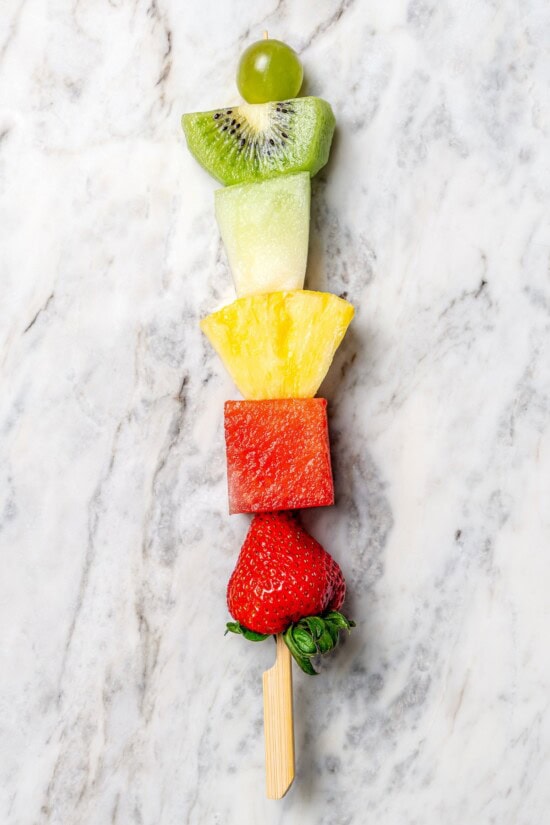 Fruit kabobs are the easiest summer dessert! Made with a rainbow of fruit threaded onto skewers, they’re perfect with a creamy fruit dip. Overhead view of single fruit skewer on marble countertop