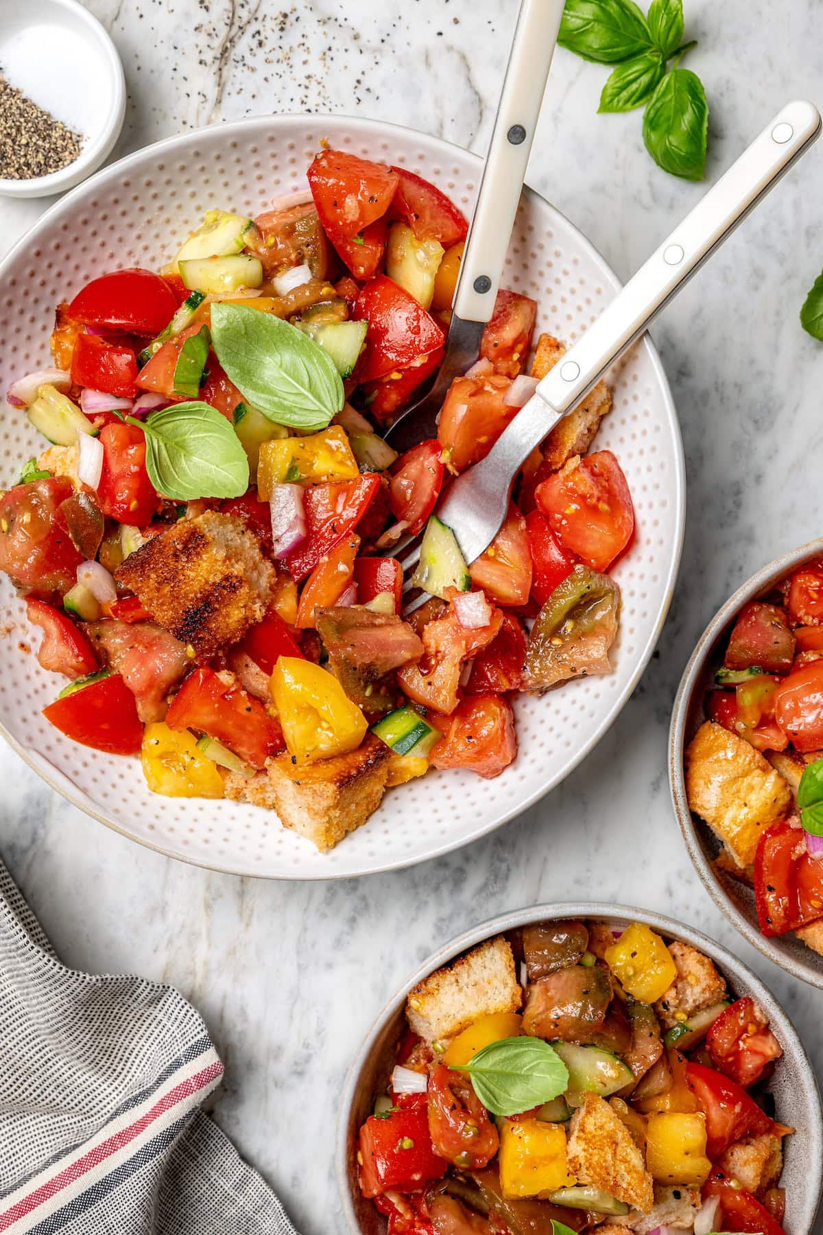 With juicy heirloom tomatoes, crisp cubes of bread, cucumber, and fresh basil, this panzanella recipe is a dish you’ll find yourself making all summer long. Serve it as a side or as a light meal. Overhead view of panzanella salad in serving bowl with fork and spoon and two smaller bowls