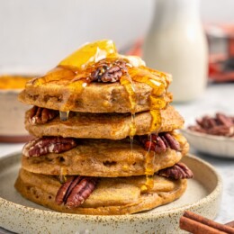 Stack of pumpkin pancakes on plate with syrup, pecans, and butter