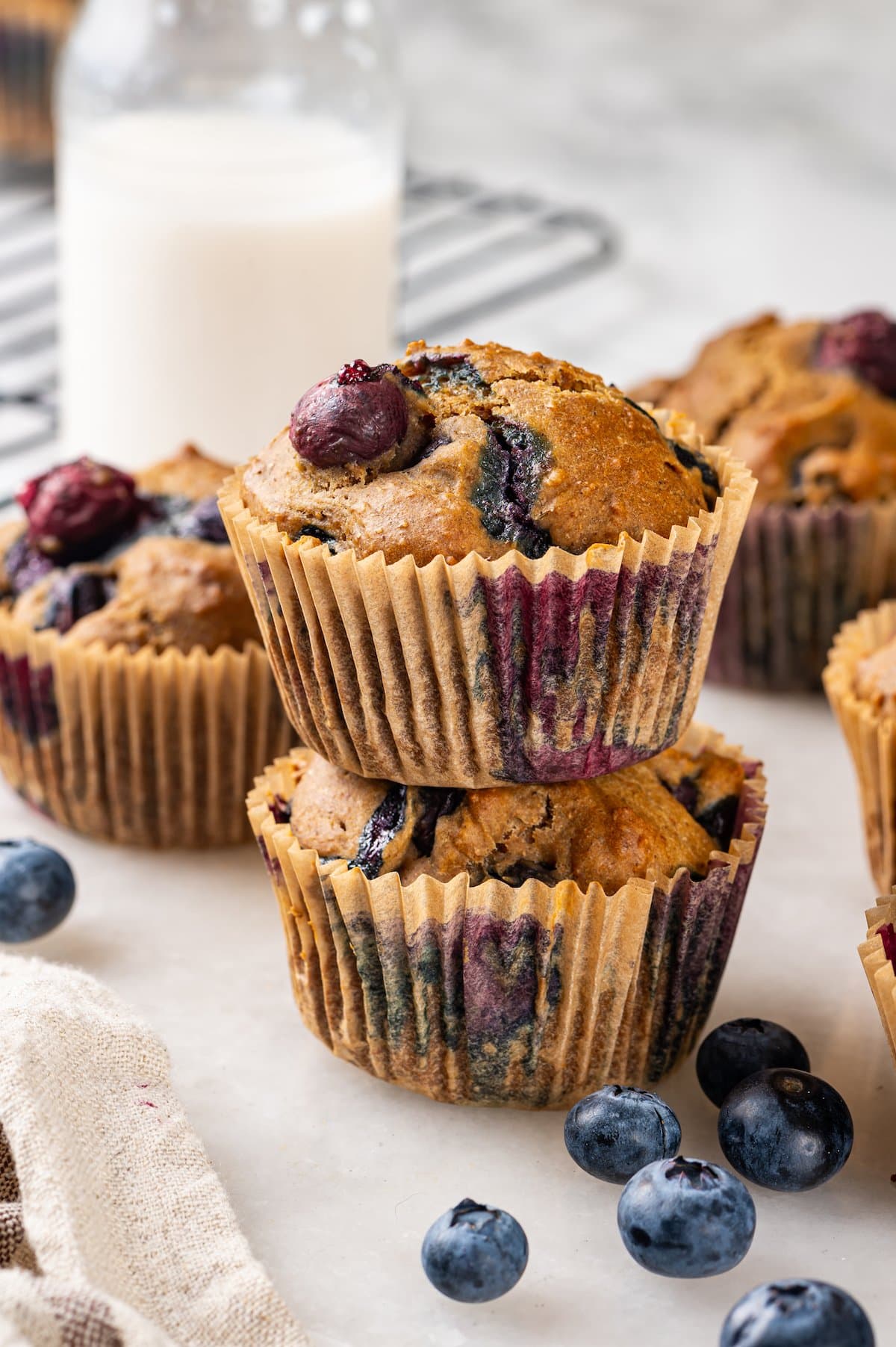 A classic blueberry muffin recipe lightened up with whole wheat flour and applesauce in place of the oil. So moist and delicious! Stack of 2 blueberry muffins with additional muffins and jug of milk in background
