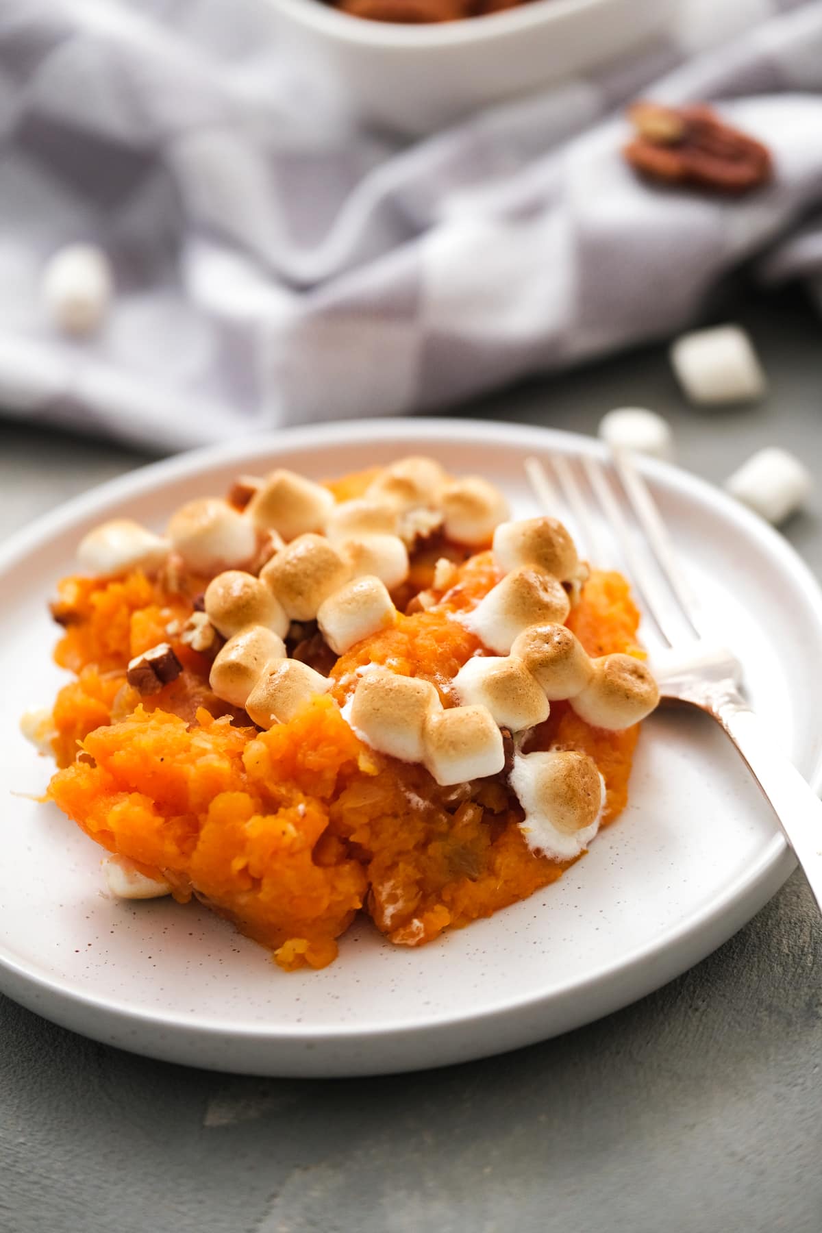 This lighter sweet potato casserole is made with golden raisins, crushed pineapple, and spices, topped with toasty marshmallows and pecans. A serving of sweet potato casserole with marshmallows on a plate next to a fork.