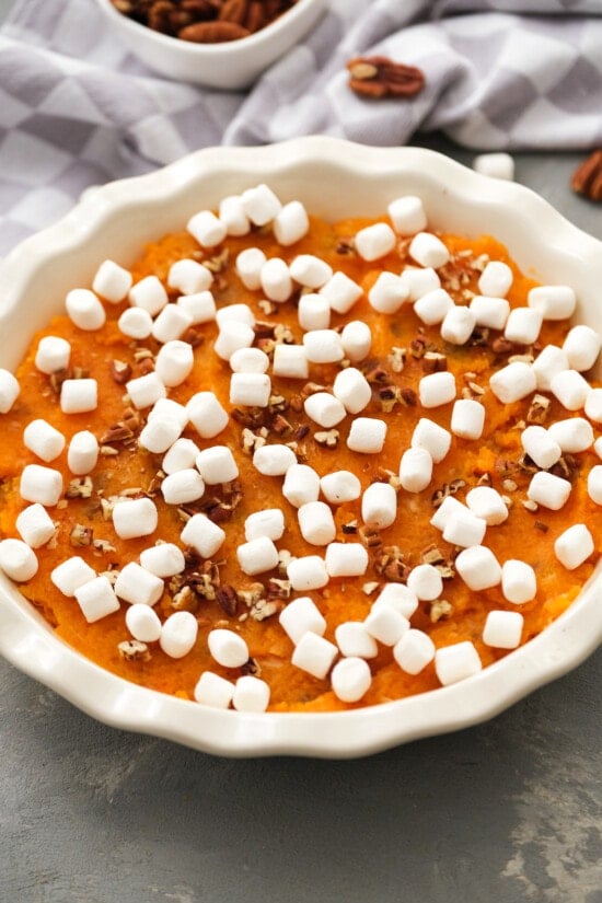 This lighter sweet potato casserole is made with golden raisins, crushed pineapple, and spices, topped with toasty marshmallows and pecans. Overhead view of an unbaked sweet potato casserole topped with mini marshmallows and chopped pecans in a round baking dish.