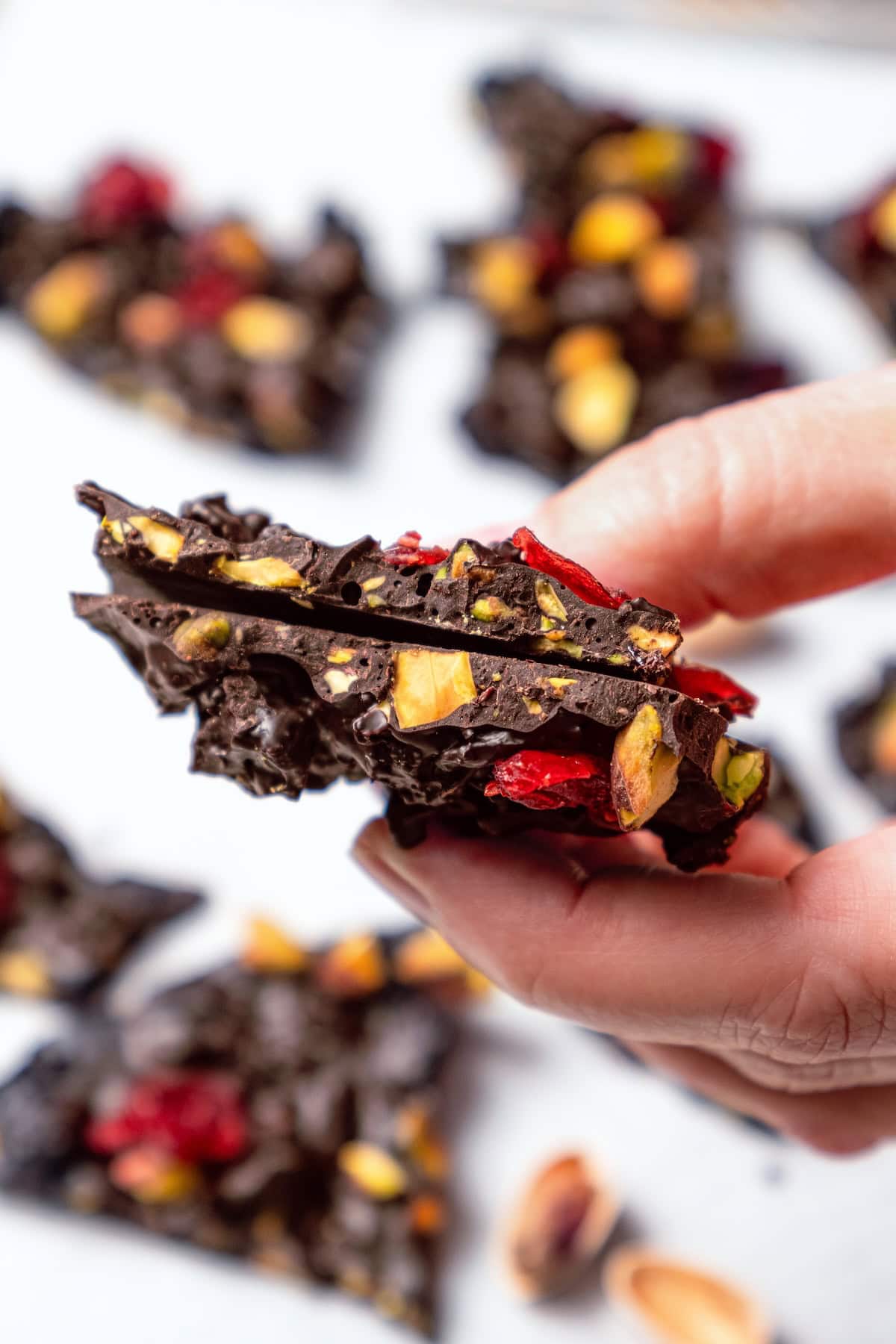 This easy Dark Chocolate Bark with Pistachios and Cranberries is festive enough for a holiday treat, but healthy enough to enjoy year round! Hand holding two pieces of dark chocolate bark with pistachios and cranberries.