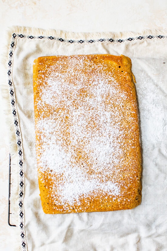 This classic pumpkin roll recipe features an irresistible pumpkin spice sponge cake with a tangy cream cheese filling. Perfect for holidays! Overhead view of pumpkin cake on a towel with powdered sugar