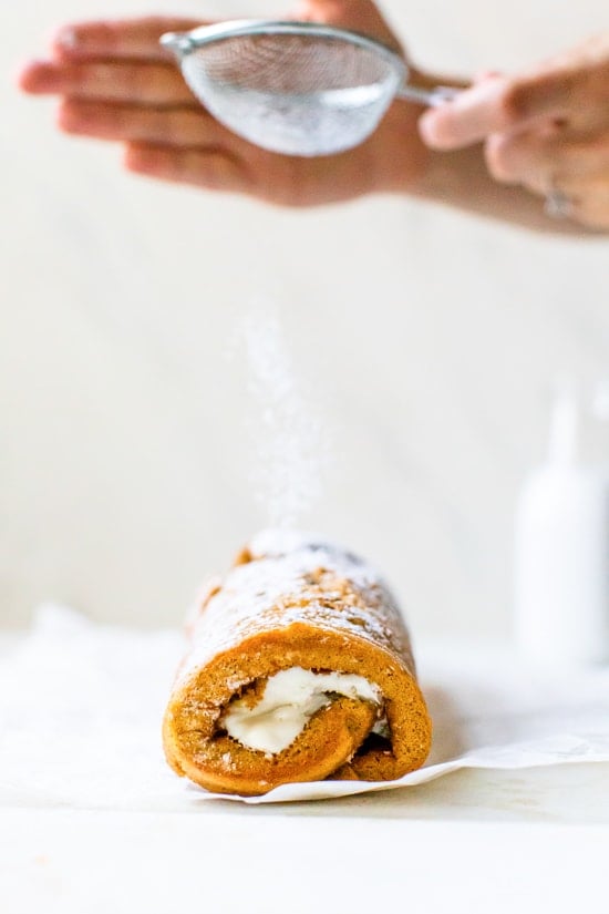 This classic pumpkin roll recipe features an irresistible pumpkin spice sponge cake with a tangy cream cheese filling. Perfect for holidays! Powdered sugar being sprinkled on a pumpkin roll