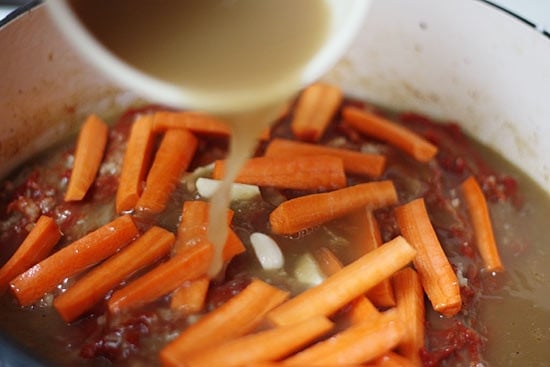 This braised brisket recipe is slow cooked in the oven with potatoes, carrots and onions. Slicing it half-way through makes it extra tender! Pouring broth into pot with carrots and brisket.
