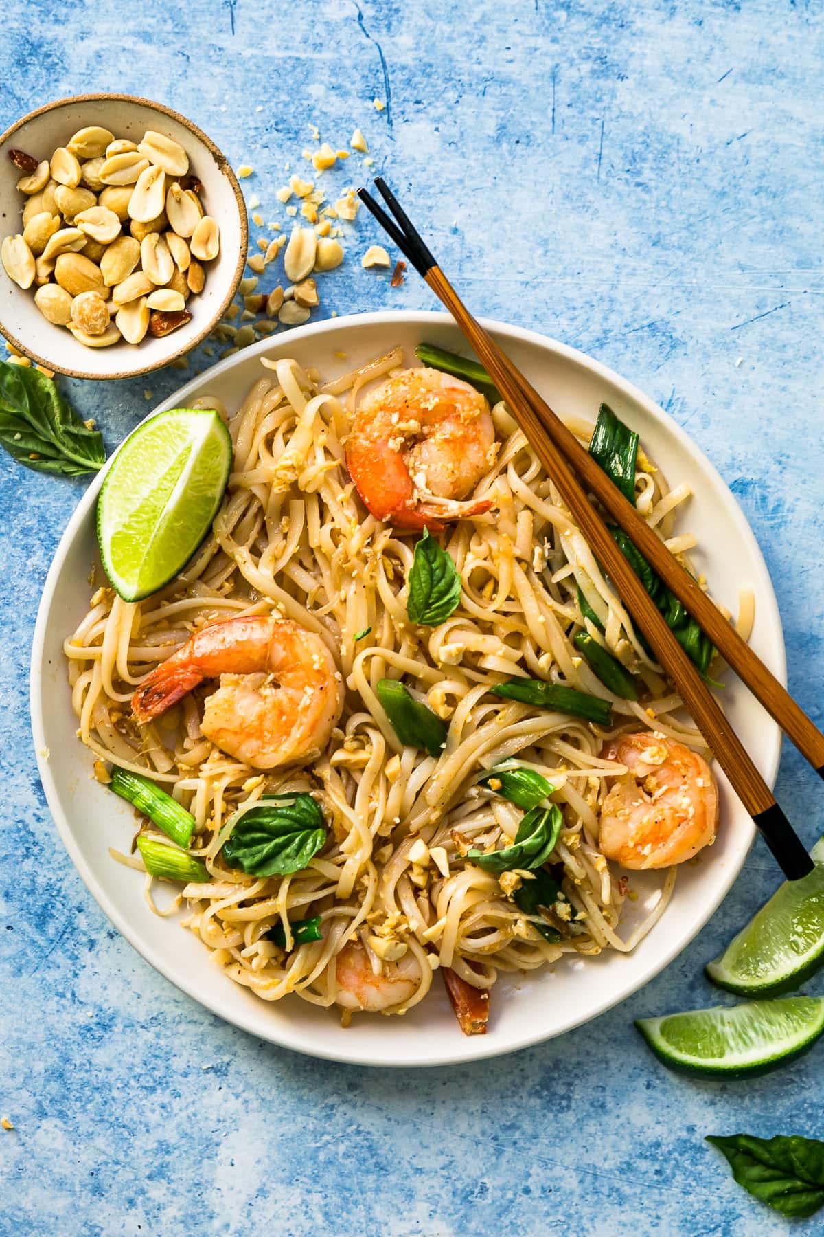 This easy homemade Pad Thai recipe is a lightened up version of the original made with shrimp, eggs, and tofu. Dinner’s done in 25 minutes! Overhead view of shrimp Pad Thai in bowl with peanuts, lime wedges, and basil for garnish