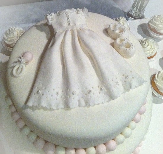 A white cake with a lacy baptism dress, baby shoes, and a rattle