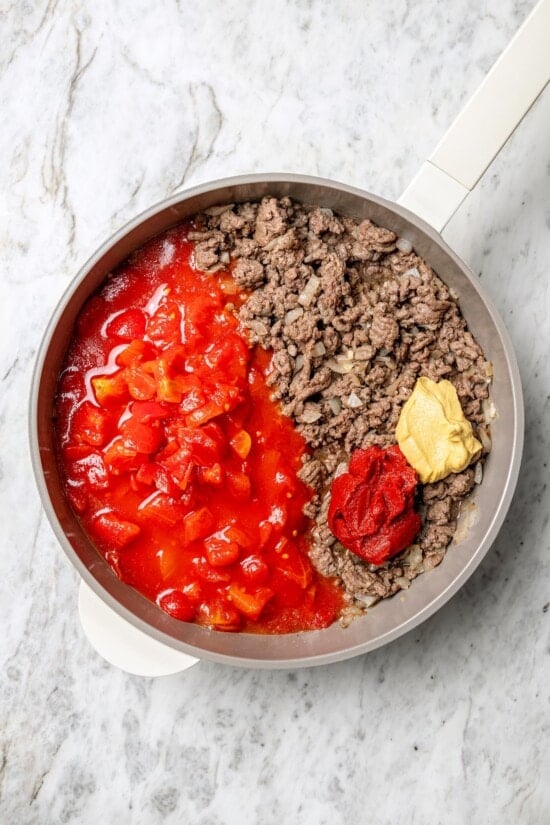 With ground beef, tomatoes, pickles, and cheese, this lightened up cheeseburger casserole recipe is kid-friendly and so delicious! Overhead view of mustard, tomato paste, and tomatoes added to skillet of cooked ground beef