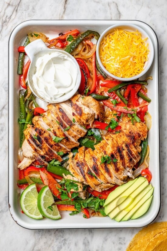 These easy chicken fajitas are made with grilled chicken breast, bell peppers and onions and served sizzling hot with tortillas and cheese! Overhead view of chicken fajitas in pan with avocado, cheese, and sour cream
