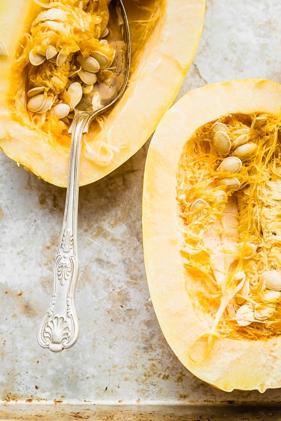 Two halves of an uncooked spaghetti squash with a spoon scooping seeds out of one half