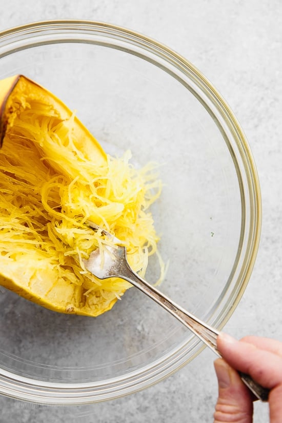 A cooked half of a spaghetti squash in a bowl with a fork removing the squash strands from the shell