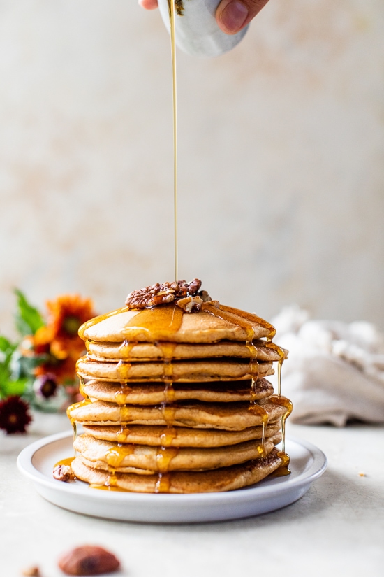 Made with whole wheat flour, pumpkin puree, pumpkin spice, and pecans, these Pumpkin Pancakes are soft, fluffy, and filled with fall flavor. Syrup being poured over a stack of pumpkin pecan pancakes