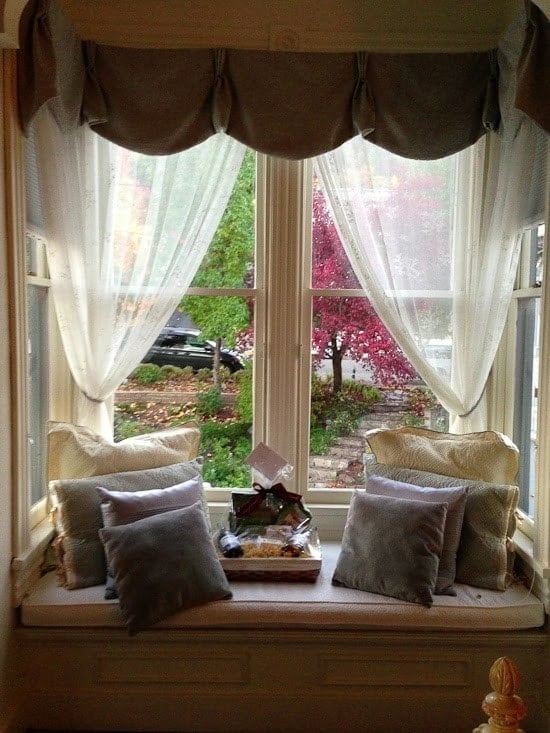 A window seat with sheer curtains, pillows and welcome basket with wine and snacks