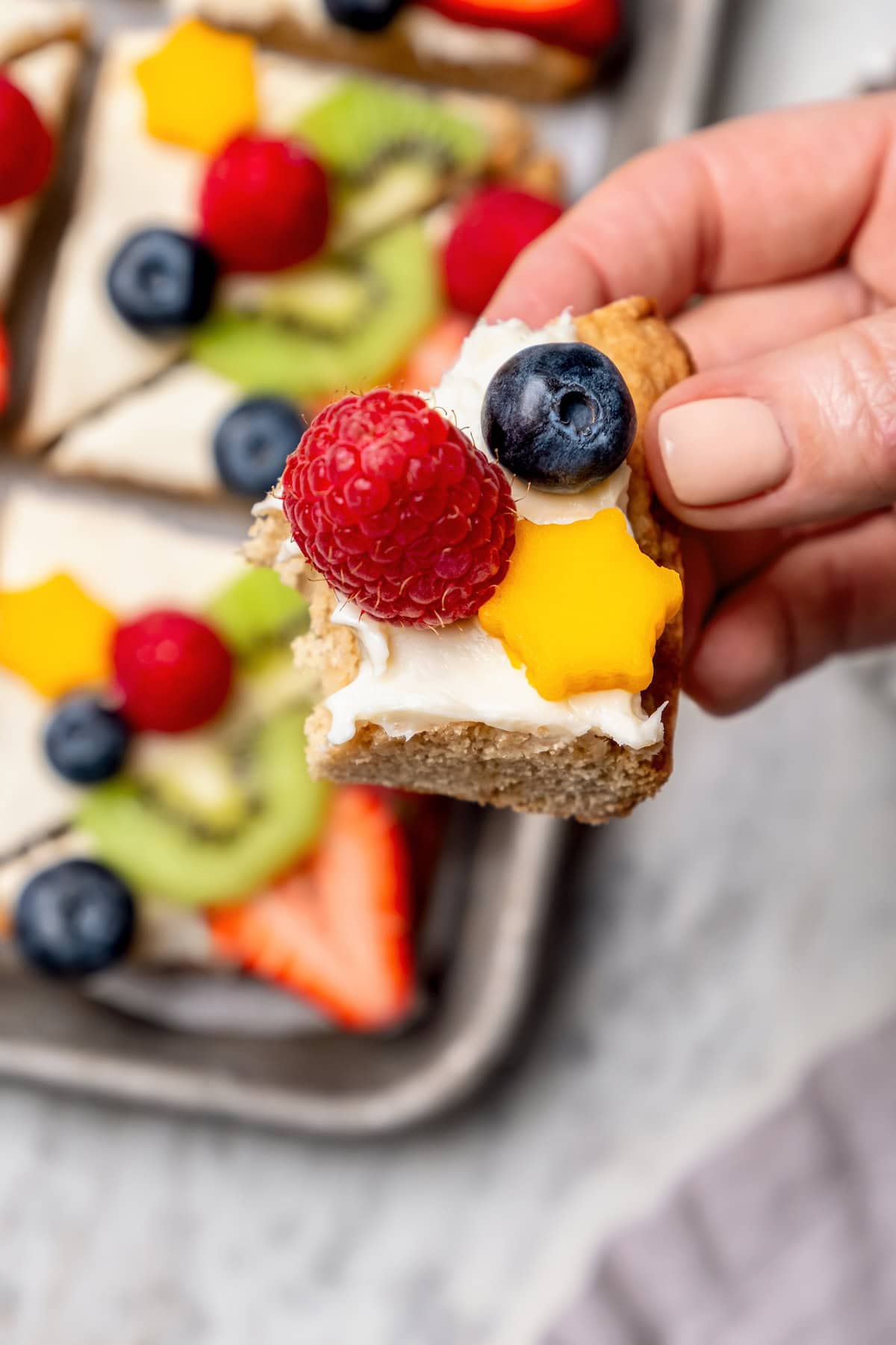 This easy fruit pizza is made with a lightened-up sugar cookie crust and topped with fluffy cream cheese frosting and colorful fresh fruit! Hand holding piece of fruit pizza