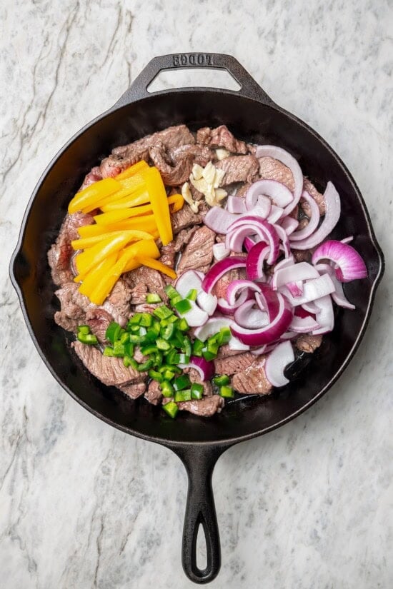 This lightened up Peruvian lomo saltado recipe is packed with flavor, with tender sirloin and veggies served alongside crispy baked fries. Overhead view of vegetables added to beef in skillet