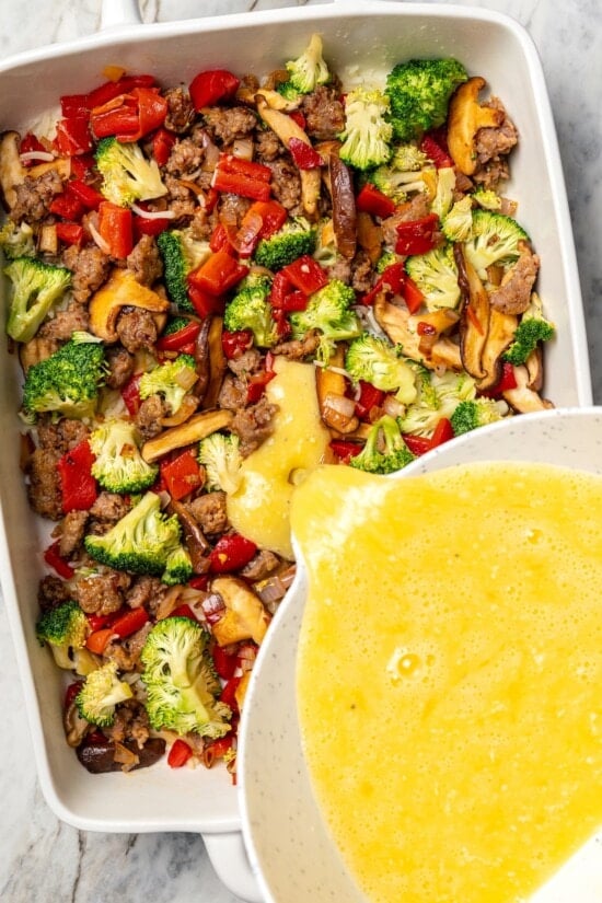 This healthy breakfast casserole recipe with sausage, cheese and veggies is hearty, flavorful, and perfect for meal prep or a weekend brunch! Pouring egg over vegetables and sausage for breakfast casserole.