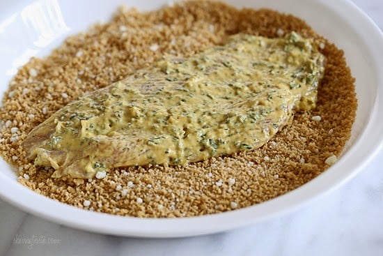 Raw chicken breast covered in dijon mustard with herbs, placed on a plate with breadcrumbs.