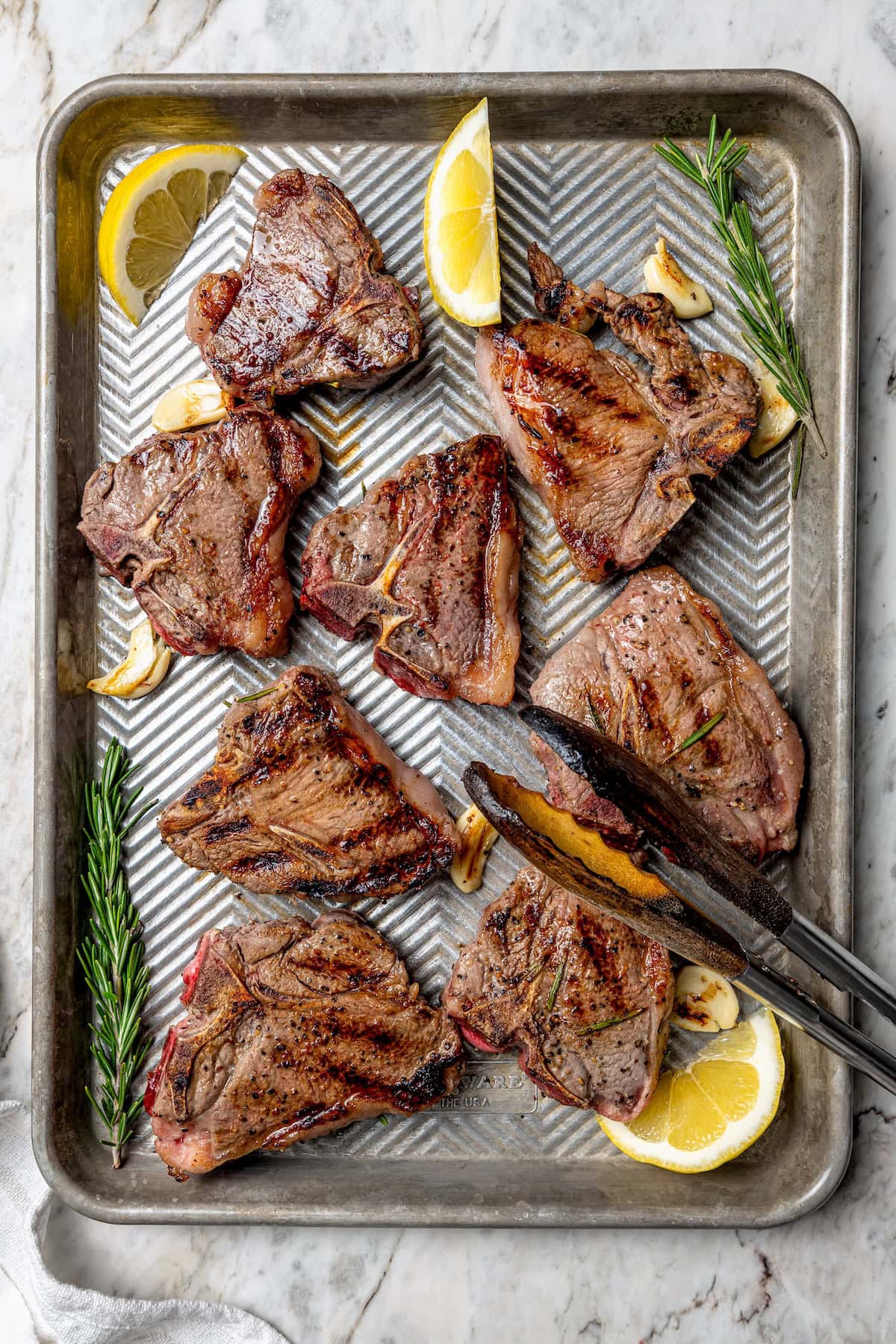 With fragrant rosemary and garlic, this easy grilled lamb chops recipe is flavorful, succulent, and one I love to cook all year long! Overhead view of grilled lamb chops on sheet pan with fresh rosemary, lemon wedges, and garlic cloves