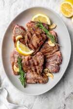Overhead view of grilled lamb chops on platter with fresh rosemary, lemon wedges, and garlic cloves