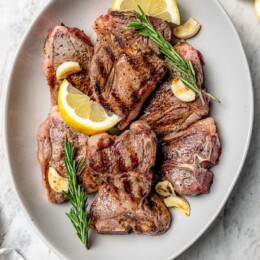 Overhead view of grilled lamb chops on platter with fresh rosemary, lemon wedges, and garlic cloves
