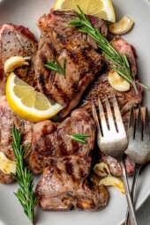 Overhead view of grilled lamb chops on platter with fresh rosemary, lemon wedges, and garlic cloves