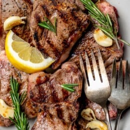 Overhead view of grilled lamb chops on platter with fresh rosemary, lemon wedges, and garlic cloves