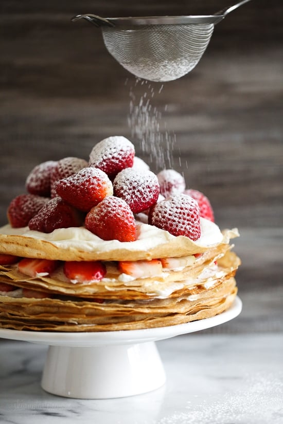 This easy strawberry crêpe cake is a light summer dessert ready to impress! A few simple swaps make it healthier than the traditional version. Dusting strawberry crepe cake on cake stand with powdered sugar