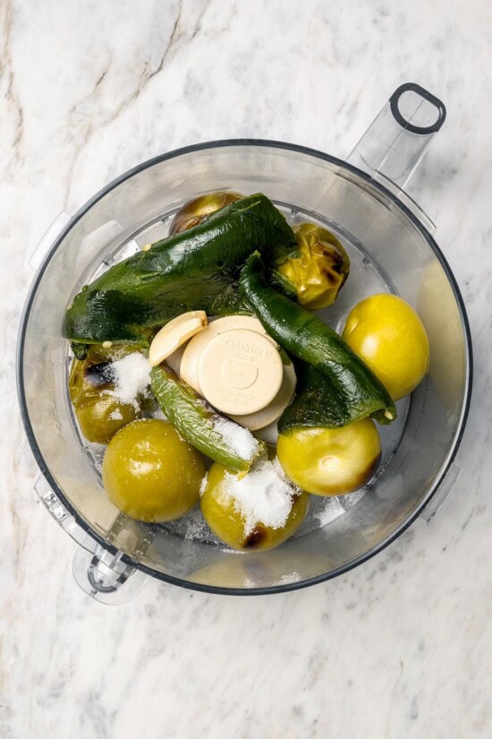 Roasting the tomatillos and peppers first makes this homemade salsa verde recipe extra delicious. Easy to make and better than store-bought! Overhead view of garlic, salt, peppers, and poblanos in food processor bowl