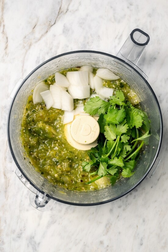 Roasting the tomatillos and peppers first makes this homemade salsa verde recipe extra delicious. Easy to make and better than store-bought! Overhead view of onion and cilantro added to salsa verde in food processor
