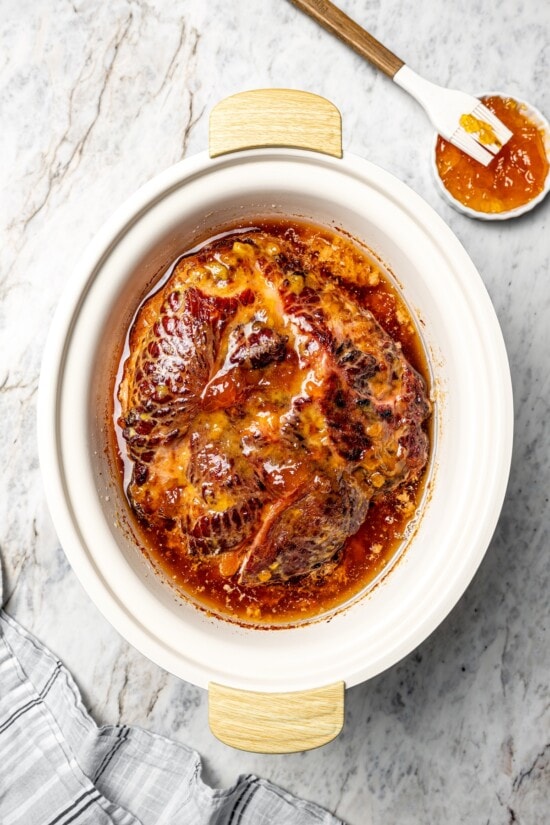 Slow Cooker Ham is the easiest main dish for the holidays! A two-ingredient glaze and a crockpot are all you need for moist, flavorful ham. Ham in slow cooker next to small bowl of apricot preserves.