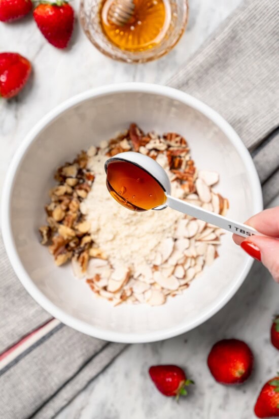 This healthy strawberry crisp recipe is perfectly portioned into individual servings, with a delicious nutty topping. So easy to make too! Drizzling honey from tablespoon into bowl of nuts and almond flour