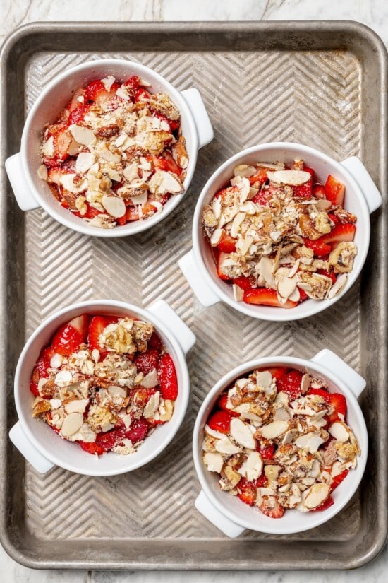 This healthy strawberry crisp recipe is perfectly portioned into individual servings, with a delicious nutty topping. So easy to make too! Overhead view of strawberry crisps in ramekins on baking sheet before baking