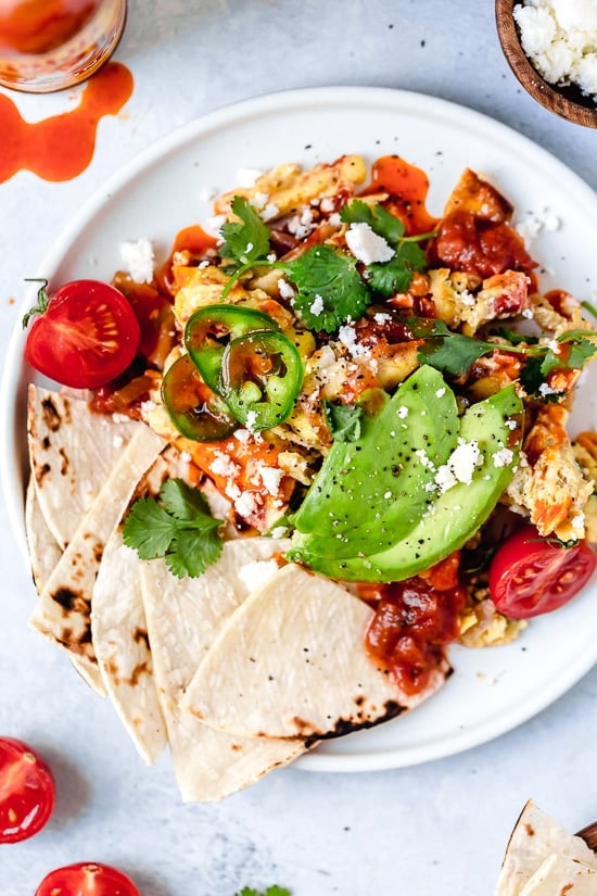 Migas are a classic Tex Mex breakfast recipe, but you can enjoy them anytime of day! Eggs, tortillas, cheese and veggies make them satisfying. Overhead view of migas on plate with tortillas and avocado