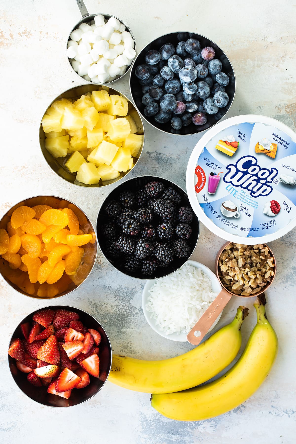 With a variety of fresh fruit, whipped cream, walnuts, and marshmallows, Ambrosia Fruit Salad is ready in minutes. A perfect summer dessert! Cool Whip, and bowls of bruit with small marshmallows.