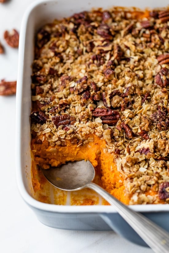 Spoon digging into sweet potato casserole with praline topping in baking dish.