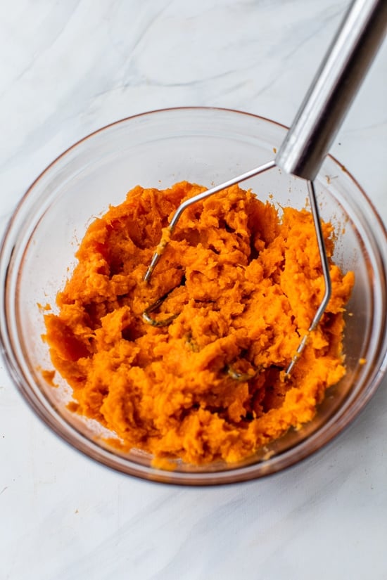 This lightened up Sweet Potato Casserole is made with roasted sweet potatoes and a praline topping for a crowd-pleasing Thanksgiving side! Potato masher mashing sweet potatoes in glass bowl.