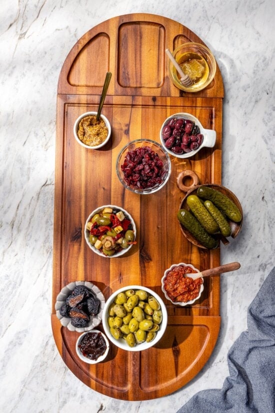 Your ultimate guide to creating a charcuterie and cheese board! I'll show you how to choose and arrange everything for a beautiful spread. Bowls of spreads, pickles, and olives on wooden charcuterie board.