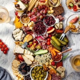 Plates surrounding large charcuterie and cheese board with fruit, pickles, olives, and spreads.