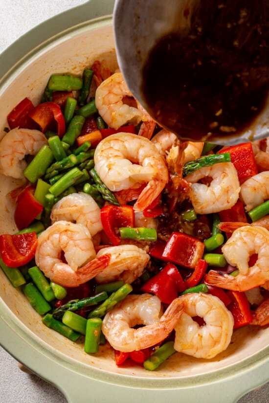 With juicy shrimp, asparagus, and red bell peppers, this Shrimp Stir Fry recipe makes a quick, easy, and healthy dinner in just 20 minutes! Stir fry sauce being poured over shrimp, asparagus, and red bell peppers
