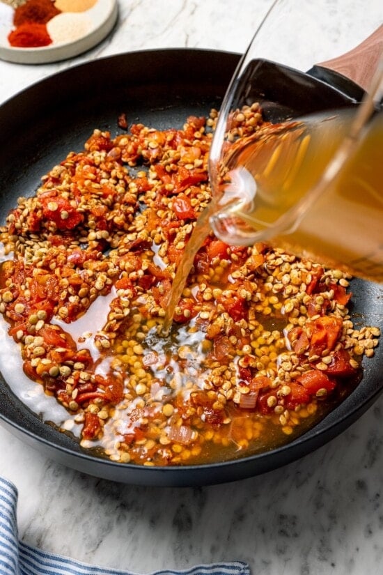 These vegan lentil tacos are easy to make, full of big Tex-Mex flavor and great for meal prep. Add your favorite toppings and dig in! Pouring broth into pan with lentils and tomatoes