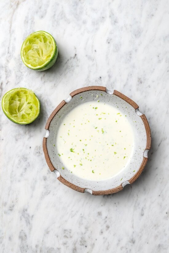 This lighter version of the classic grilled Mexican street corn recipe has all the flavor of the original, with mayo, cotija, lime and Tajín! Overhead view of lime mayo in bowl set next to 2 juiced lime halves