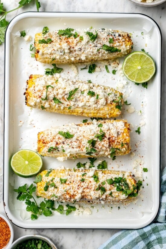 This lighter version of the classic grilled Mexican street corn recipe has all the flavor of the original, with mayo, cotija, lime and Tajín! Overhead view of 4 ears of Mexican street corn in pan with lime halves and cilantro
